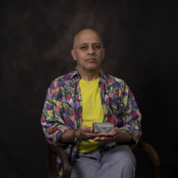 Portrait of Ali wearing a floral shirt and yellow t-shirt, seated and holding a decorative mina-kari box – part of the Faces of Resilience series by Masoud Teimory