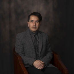 Portrait of Behnam in a dark grey jacket and buttoned shirt, seated against a softly lit painterly backdrop – part of the Faces of Resilience series by Masoud Teimory