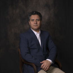 Portrait of Farnood in a dark blue jacket and open-collar white shirt, seated against a painterly brown backdrop – part of the Faces of Resilience series by Masoud Teimory