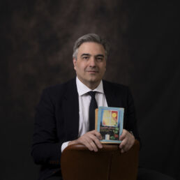 Portrait of Hatef in a black suit and tie, seated and holding a Persian Nowruz book – part of the Faces of Resilience series by Masoud Teimory