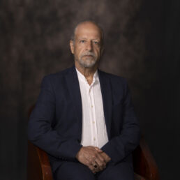 Portrait of Hesam in a dark blue jacket and white shirt, seated with hands clasped – part of the Faces of Resilience series by Masoud Teimory