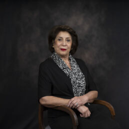 Portrait of Manoush in a black outfit and patterned scarf, seated with a warm expression – part of the Faces of Resilience series by Masoud Teimory