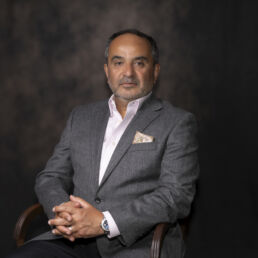 Portrait of Mehran in a grey suit with pink shirt and patterned pocket square, seated against a painterly backdrop – part of the Faces of Resilience series by Masoud Teimory