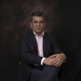 Portrait of Peyman seated in a dark jacket and pink shirt, against a painterly dark backdrop – part of the Faces of Resilience series by Masoud Teimory