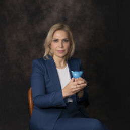 Portrait of Sanaz in a blue suit, seated against a dark backdrop, holding a turquoise ceramic bird – part of the Faces of Resilience series by Masoud Teimory