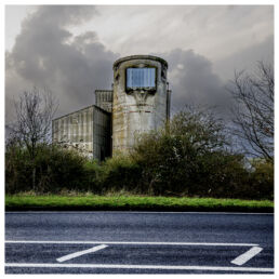 Derelict concrete building with a square tower viewed from across a wet road under a stormy sky