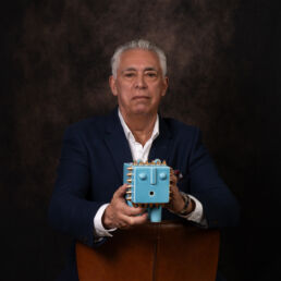 Portrait of Fash in a navy blue suit, holding a turquoise ceramic sculpture with gold spikes – part of the Faces of Resilience series by Masoud Teimory